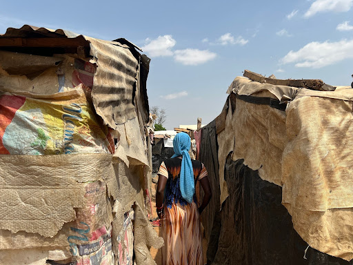 Person in a colorful dress and blue headscarf walks through narrow pathways between makeshift shelters under a clear sky.
